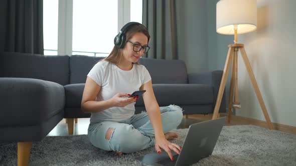 Casually Dressed Woman with Headphones is Sitting on Carpet with Laptop and Smartphone and Working