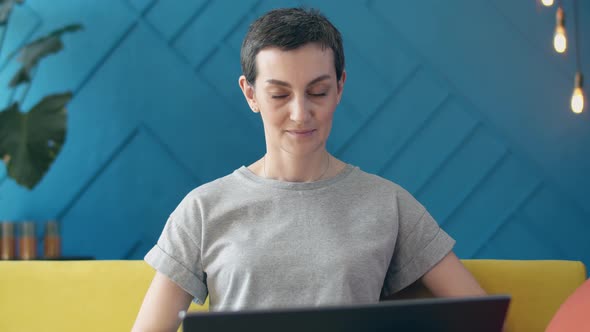 Woman with Short Hair Writes a Text on a Laptop Sitting on Couch in the Room and Shrugs Spreading