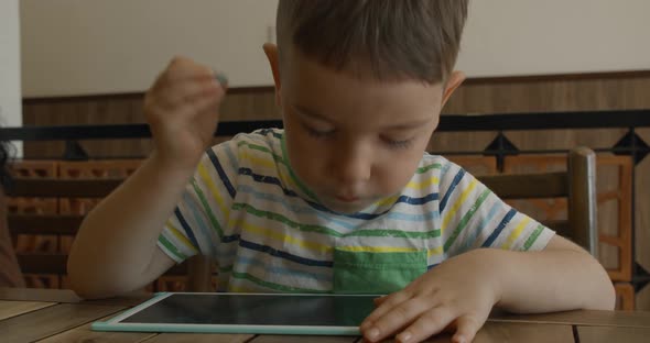 A Portrait of a Child Sits at a Table at Home and Draws on His Tablet with a Special Pen a