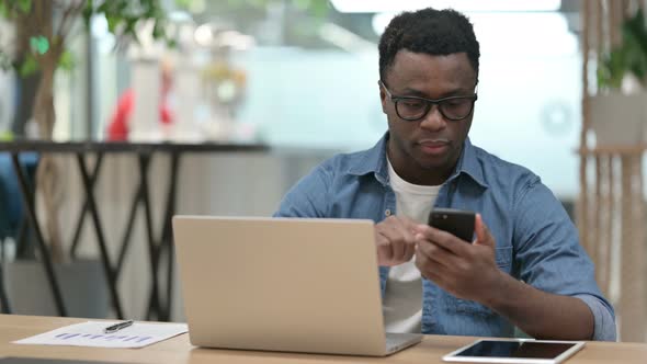 Young African Man Using Smartphone While Working on Laptop
