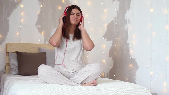 A Beautiful Girl in Pajamas Listening to Music on Headphones While Sitting on a Bed in a Good Mood