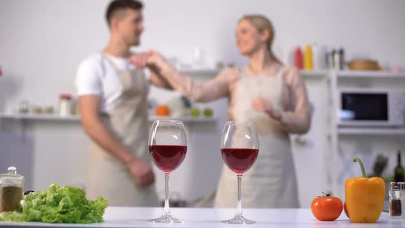 Glasses of Red Wine on Table, Romantic Couple Dancing on Background in Kitchen