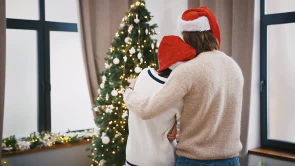 Unrecognizable Young Man and Woman in Santa Hats are Hugging Standing at Home Against Decorated