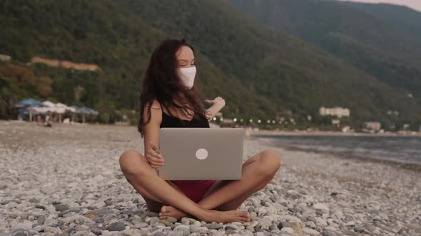 Young Woman in Medical Mask Sitting on the Beach and Talking on Video Conference with Friends