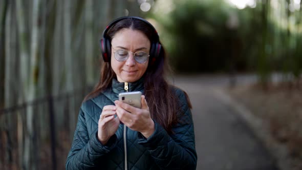 Carefree Brunette Woman with Cell Phone and Wireless Headphones is Enjoying Music in Park