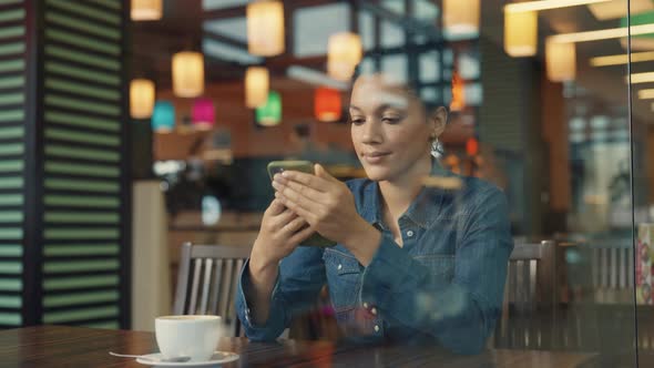 View Through Glass Showcase of Young African American Woman Texting on Phone and Enjoying Coffee in