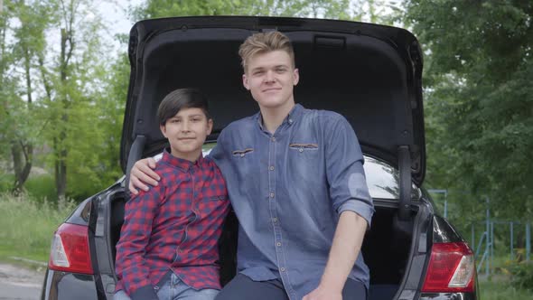 Portrait Young Father and His Son Sitting on the Back of the Car Outdoors. The Man Hugging Boy