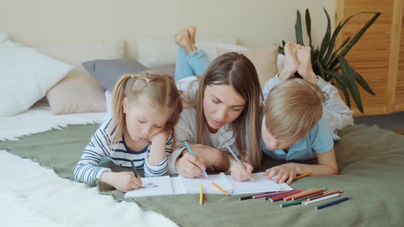 Family Leisure Mom and Kids Lay on Bed and Draw Pictures on Paper in Apartment