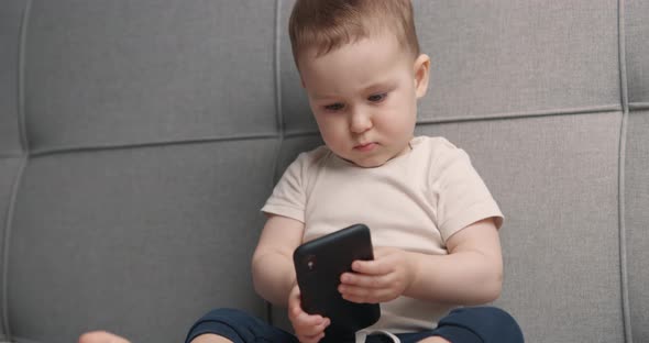 Little Boy Sitting on Bed with Mobile Phone in Hands and Playing