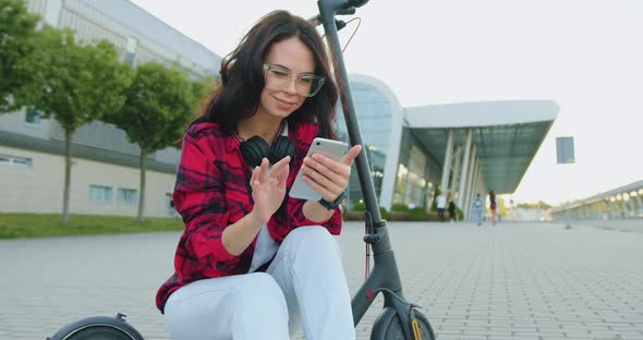 Woman with Headphones which Reviewing Funny Videos on Mobile while Sitting on Electric Scooter