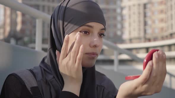 Close-up of Charming Young Middle Eastern Woman in Hijab Applying Face Powder Outdoors. Portrait 