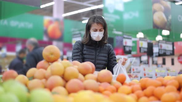 Young Woman in a Protective Mask in the Supermarket Chooses Fruit and Puts It in a Plastic Bag