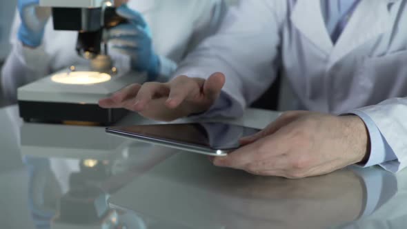 Male Scientist Scrolling Tablet Screen, Colleague Working With Microscope Nearby