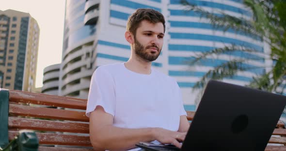 A Young Man in the Summer of Palm Trees Sitting with a Laptop and Typing on the Keyboard