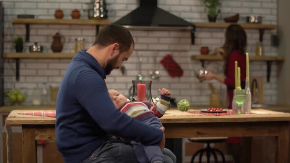 Bearded Father Playing with Baby Little Son, Sitting at the Table While His Wife Cooking Dinner