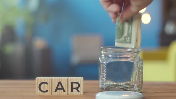 Car is word written on wooden cubes lying on table. Hand puts money in nearby jar.