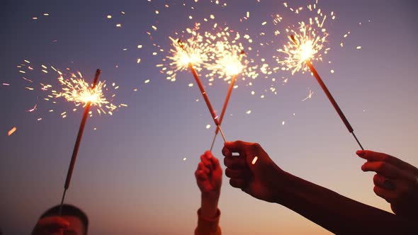 Closeup of hands holding sparklers, happy friends are waving bengal fireworks background