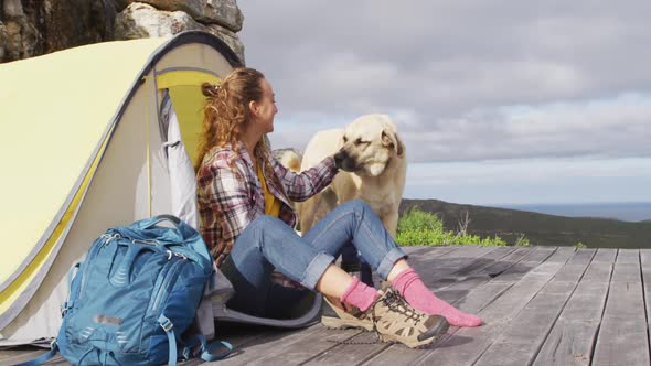 Happy caucasian woman camping sitting outside tent with pet dog, putting on boots on mountainside