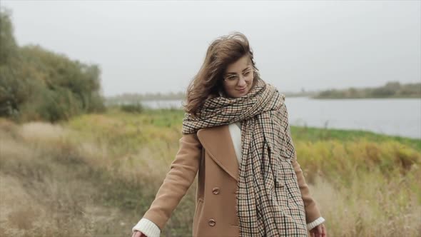 An Attractive Young Woman Walks Along the Path Along the River Wrapped in a Scarf and Smiling