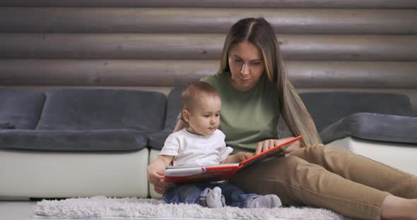 Young Mother and Her Baby Son Play with Book While Sitting on Floor at Home