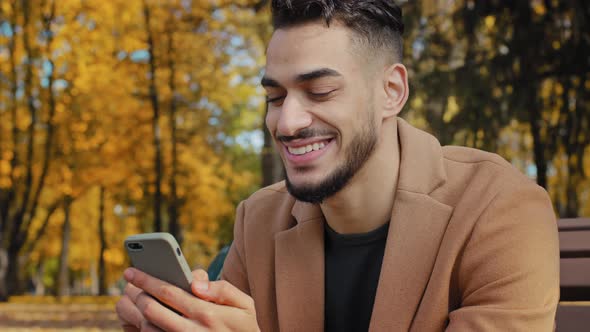 Close Up Young Hispanic Guy Sitting on Bench in Autumn Park Bearded Man Holding Mobile Phone Makes