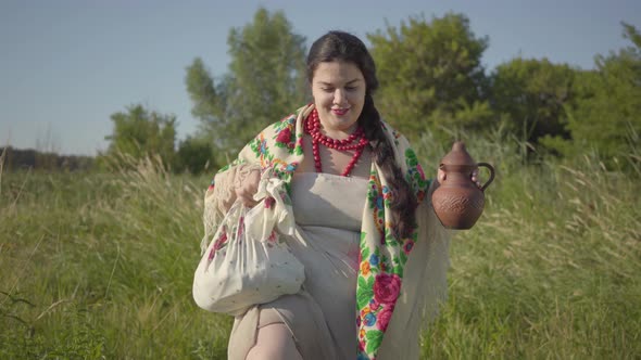 Beautiful Overweight Woman Walking in Wild High Grass with the Earthen Jar in the Green Summer Field
