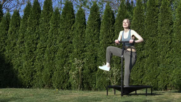 girl does Jumping fitness on trampoline in outdoor on green grass and trees on background