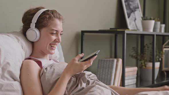 Young Woman in Headphones Playing with Dog in Bed