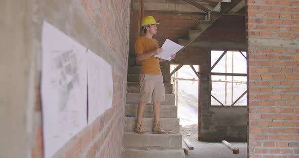 Male Building Designers Wearing Helmets In Unfinished Brick House Looking At Blueprint