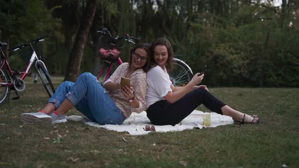 Two Young Women Sitting Back To Back at the Park and Taking a Video Call