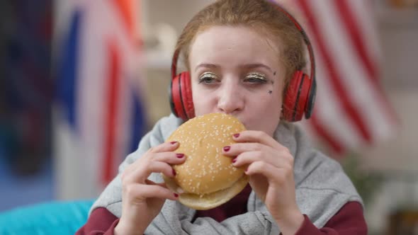 Portrait of Caucasian Hipster Woman Eating Delicious Sandwich Enjoying Music in Headphones at Home