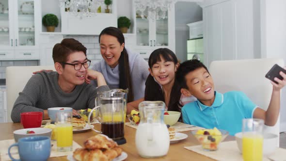 Happy asian parents in kitchen having breakfast with son and daughter, son taking group selfie