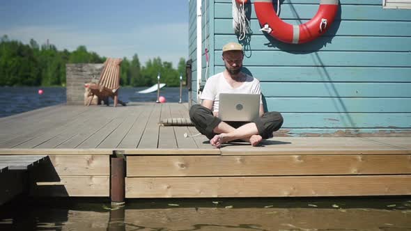 Young American Man is Working with Laptop While Sitting on Lake Pier on Summer Day Spbd