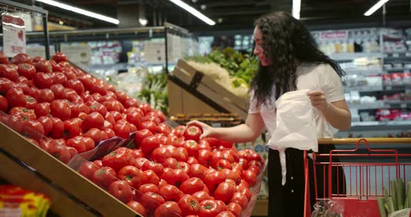 Young Woman in the Grocery Section of a Supermarket Picks Up a Bag and Puts Tomatoes in a Basket to