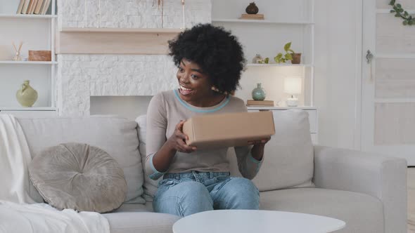 Smiling Happy African Young Woman Customer Sit on Sofa Shakes Parcel Carton Box Satisfied with