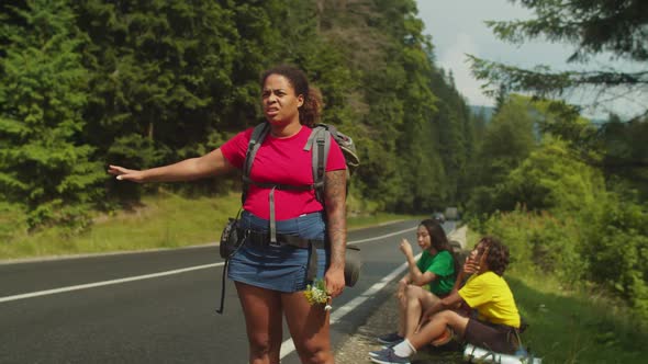 Tired African American Female Backpackers Hitchhiking on Highway During Mountain Hiking