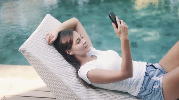 Woman Lies on Sunbed Near Pool and Uses Phone for Work Types Message