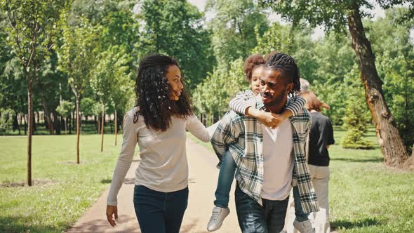 Cute African Ethnicity Girl Sits Piggyback on Her Father's Back As the Whole Family Walks Through