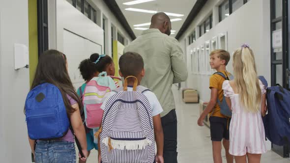 Video of back of african american male teacher and diverse pupils walking at school hall