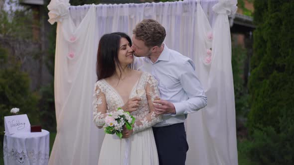 Excited Caucasian Groom Kissing Happy Middle Eastern Bride Standing at Wedding Altar with Champagne