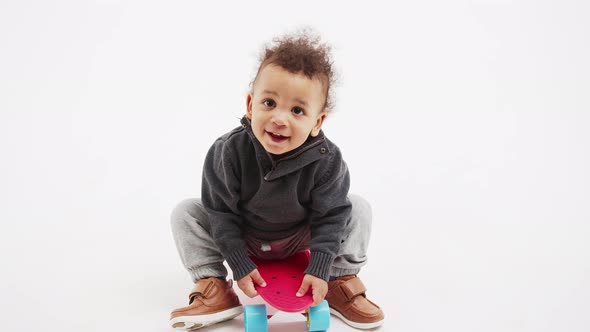 Cute Curly Toddler Sitting on the Skateboard and Looking Into the Camera Studio Shot Full Shot White