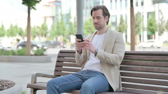 Young Man Browsing Internet on Smartphone While Sitting on Bench