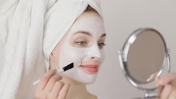 Smiling Young Charming Woman with Towel on Head Applying White Cosmetic Clay Mask on Face Using Make