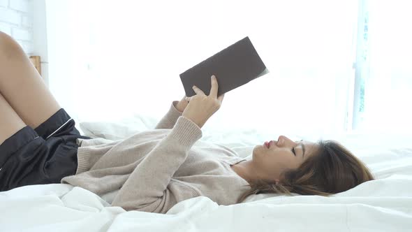 sian woman enjoying lying on the bed reading book pleasure in casual clothing at home.