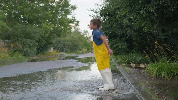 Children Holiday, Cheerful Kid in Rubber Boots and Overalls Have Fun Jumping From Curb Into Puddle