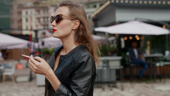 a Blonde with Bright Lipstick and Sunglasses is Walking Along a City Street with a Cigarette in Hand