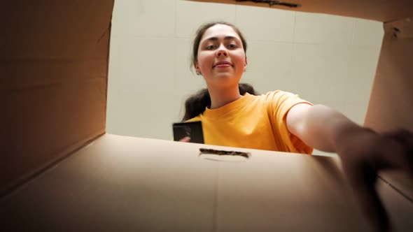 Woman chatting online on the cellphone, opens a cardboard box and takes out book