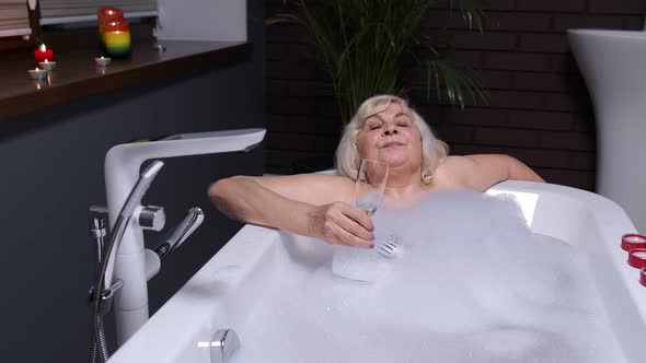 Senior Woman Grandmother Is Taking Foamy Bath, Drinking Champagne in Luxury Bathroom with Candles