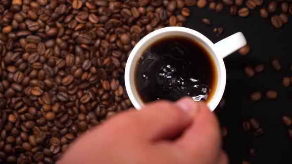 Top View of Person Hand Stirring Coffee in White Cup with Spoon on Black Table with Coffee Beans on