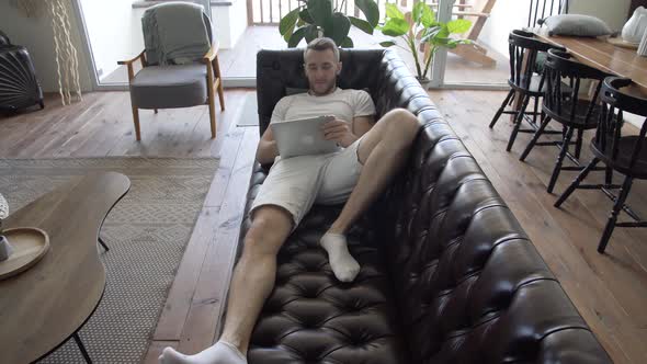 A man with a tablet in his hands is relaxing at home on the couch